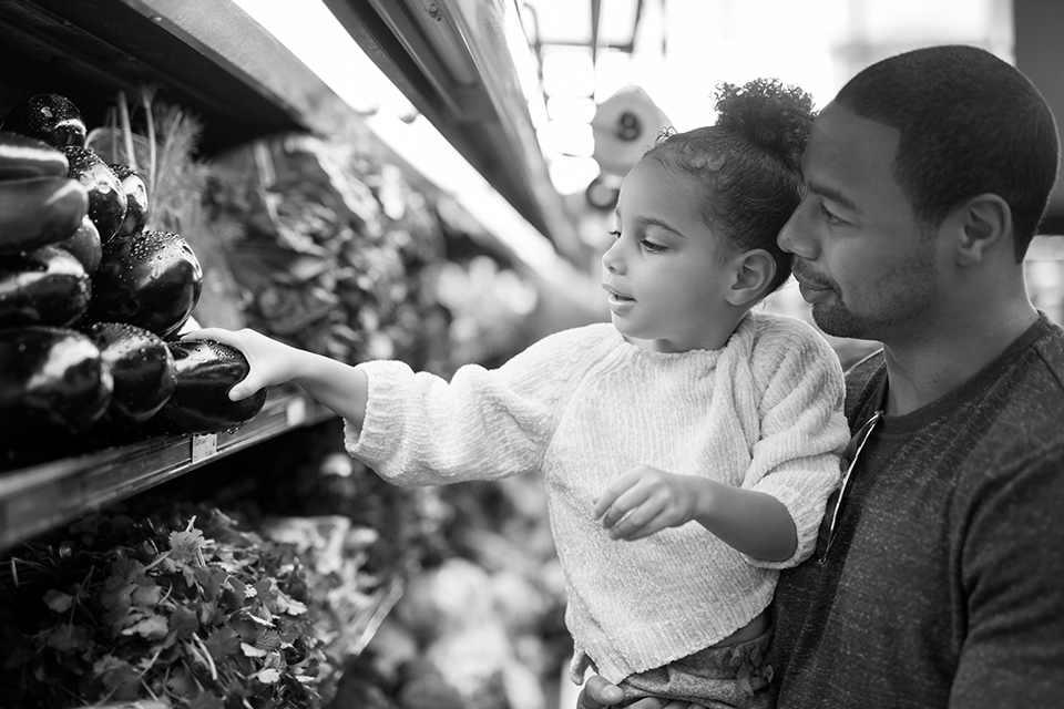 A child picking produce at a grocery store while being held by her father.